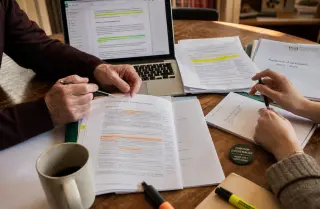 Two people in a cozy office, their hands actively engaging over annotated reports and statutes, surrounded by documents and a laptop, sunlit with a warm and authentic atmosphere.