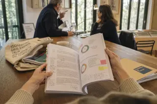 Hands examining a annotated foundation annual report on a wooden table, with a candid background scene of two people in serious discussion in a softly lit, professional office.