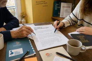 Two pairs of hands, one older and one younger, collaboratively annotating a philanthropy report at a sunlit wooden table in a warm French coworking space, surrounded by pens, coffee cups, and association folders.