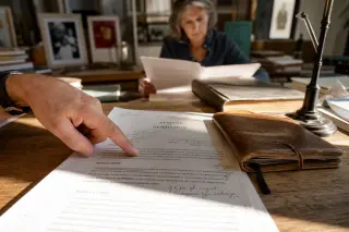 Thoughtful scene in a small foundation's office with papers and a notebook, a hand gesturing over a charter, and a woman intensely reading in soft, late afternoon light.
