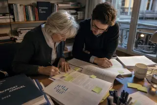 Two people, absorbed in annotating a detailed impact report at a cluttered foundation office desk, soft natural light, candid scene of authentic engagement